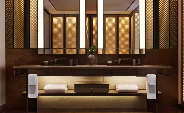 Banheiro da Suíte Deluxe com espelho iluminado e acabamentos em tons quentes no Aman New York.