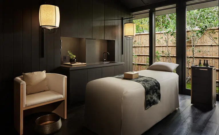 Spa treatment room at Aman Kyoto resort with serene bed, cream armchair, and garden views through large windows.
