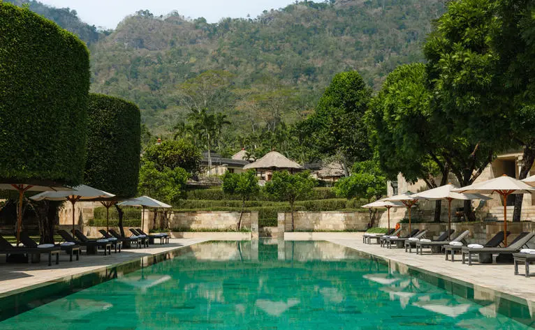 Amanjiwo's main swimming pool with emerald water, surrounded by sun loungers and cream umbrellas, set against forested hillside.