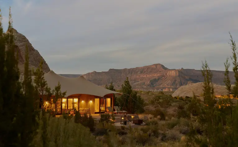 Amangiri resort at dusk, with warm-lit buildings set against desert mountains and sparse vegetation.