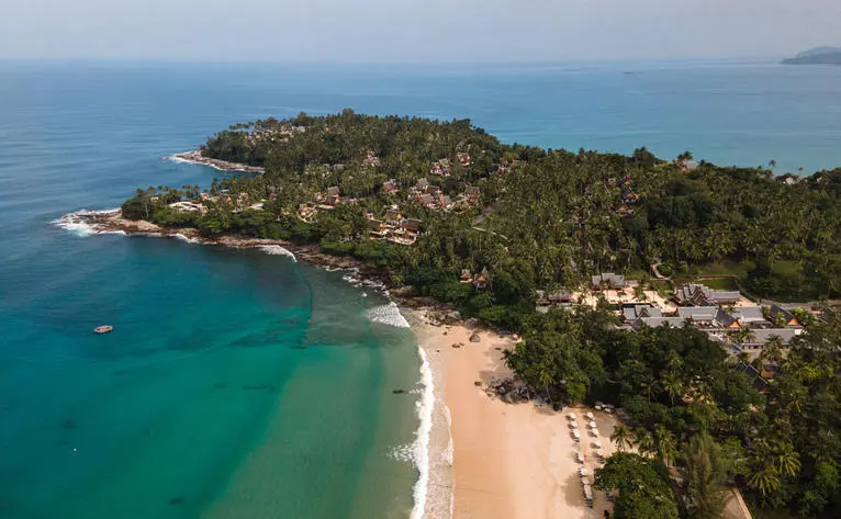 Luftaufnahme des Amanpuri-Resorts in Thailand mit Strand, türkisblauem Meer und bewaldeten Hügeln.