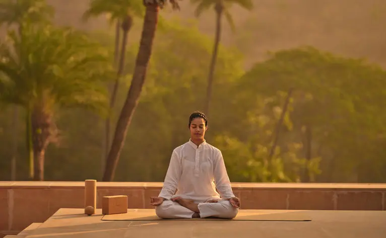 Woman meditating on terrace at Amanbagh at sunrise, overlooking palm trees and hills.