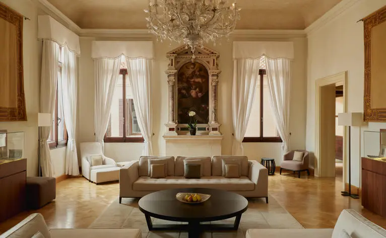 Aman Venice's Coccinas apartment living area with symmetrical seating, chandelier, and tall windows overlooking the canal.