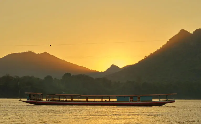 Wooden boat on the Mekong River at sunset, mountains silhouetted against golden sky at Amantaka.