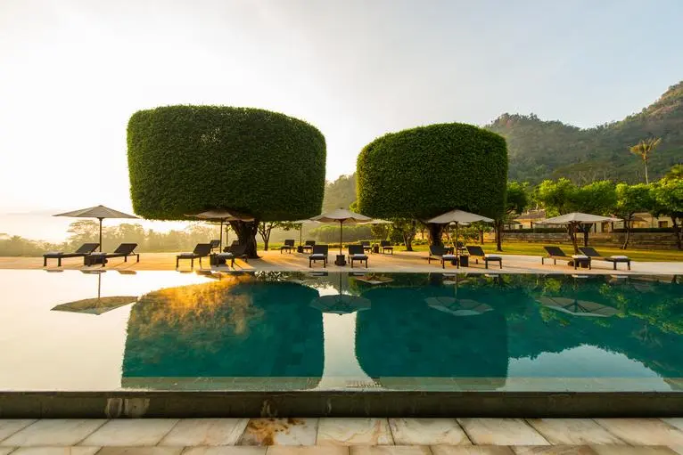 Kolam renang utama Amanjiwo dengan air biru jernih, dikelilingi pohon topiaries dan pegunungan di latar belakang.