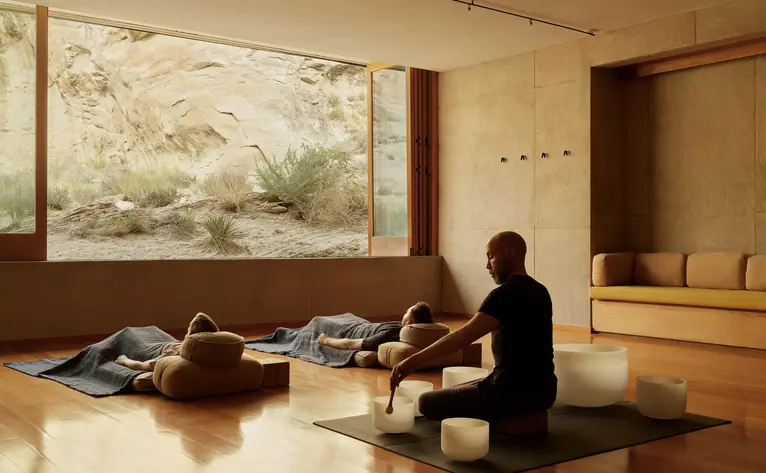 A woman sits cross-legged on a mat at Amangiri during a sound bowl wellness experience, with a large window overlooking desert landscape.
