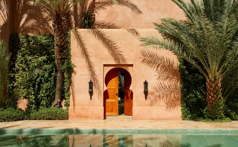 Moroccan architectural detail at Amanjena showing terracotta archway beside reflecting pool and palm trees.