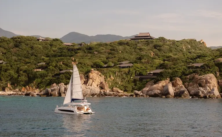 Catamaran sailing off the coastline at Amanoi resort, Vietnam, with lush forested hills and rocky shores.