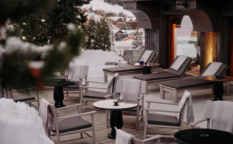 Terrace seating at Aman Le Mélézin with snow-covered chairs and evergreen branches in soft winter light.