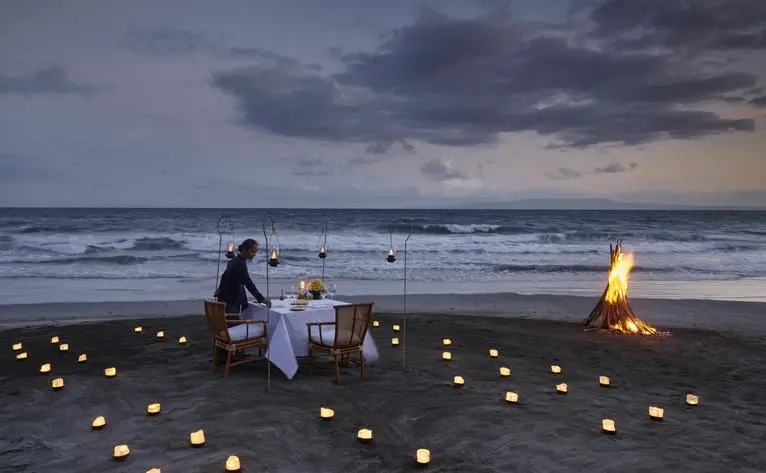 Couple dining by candlelight on Amankila's beach at dusk, with lanterns glowing along the shoreline.