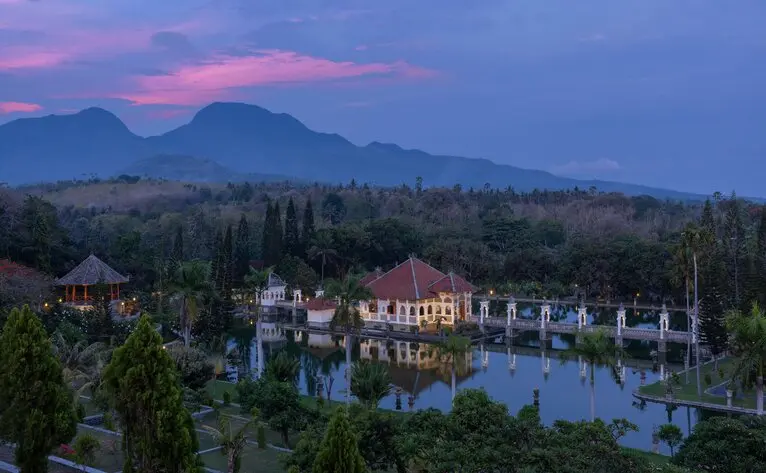 Amankila at dusk with the resort's illuminated buildings reflected in water, Mount Agung rising beyond the Balinese landscape.