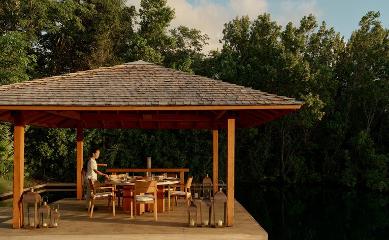 Wooden pavilion with red roof at Amanyara, furnished with dining table and chairs surrounded by tropical vegetation.