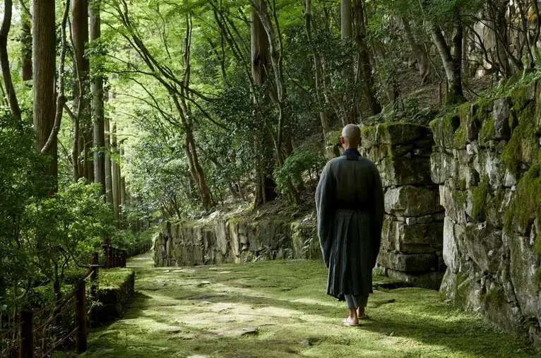 Figure in traditional black robes walking through a serene stone-lined pathway at Aman Kyoto resort, filtered sunlight through lush greenery.