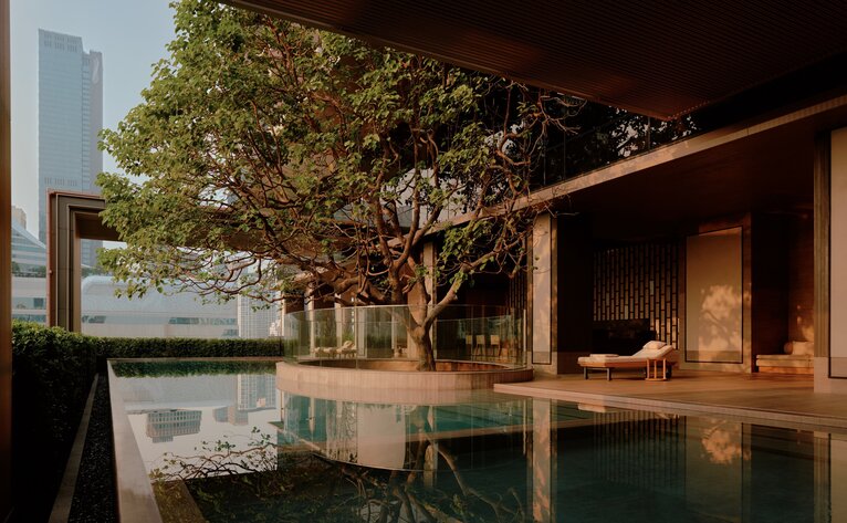 Swimming pool at Aman Nai Lert Bangkok with mature tree and wooden pavilion at dusk.