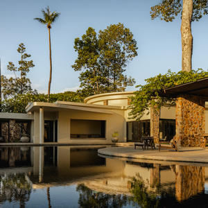 Amansara's exterior reflected in still water, with palm trees and stone architecture framed by clear sky.