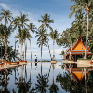 Amanpuri's infinity pool reflecting palm trees and golden-roofed pavilion under clear sky.