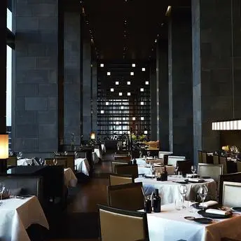 安缦东京餐厅内部，宽敞高挑的用餐空间，餐桌置于石柱之间，温暖灯光映照。