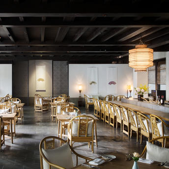 Amanfayun's dining venue with wooden tables, rattan chairs, and pendant lighting beneath dark wooden beams.