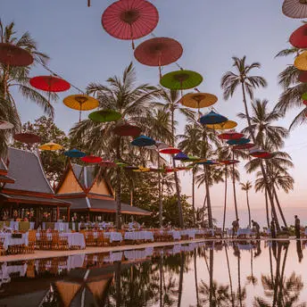 Red umbrellas suspended above a reflection pool at Amanpuri at dusk, with palm trees and pavilions beyond.