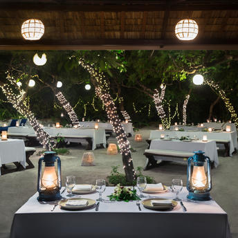 Outdoor dining table at Amanpulo with place settings and lanterns beneath illuminated trees at dusk.