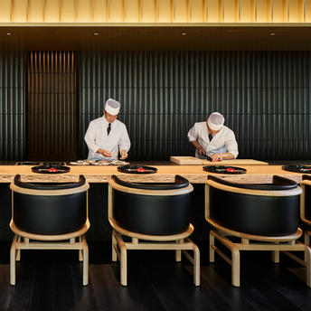 Chef's counter at Aman Kyoto resort, two chefs preparing meals behind wooden slatted counter with curved wooden chairs.