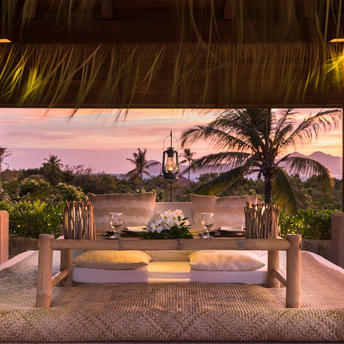 Sala dinner setup at Amanpulo with wooden pillars, tropical plants, and ocean view at sunset.