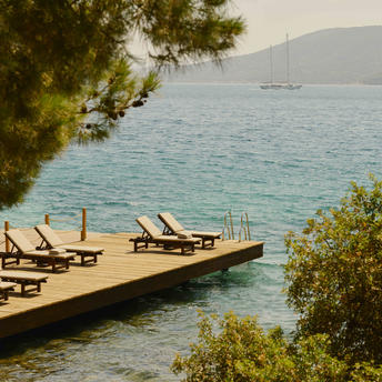 Wooden jetty with sunloungers extending into turquoise water at Amanruya, framed by pine trees.
