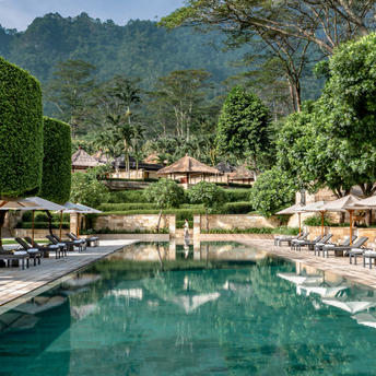 Amanpulo's main pool surrounded by sun loungers and umbrellas, with verdant hills beyond.