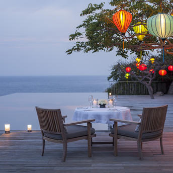Two chairs and a table on a clifftop deck at Amanoi, Vietnam, overlooking calm waters at dusk with lanterns overhead.