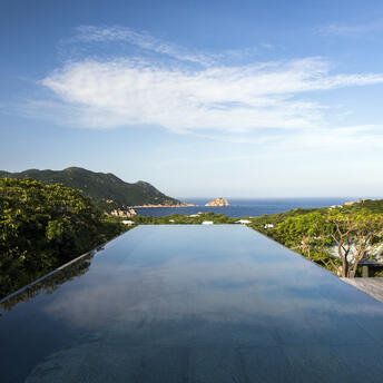 Amanoi residence overlooks a tranquil lagoon with forested hillsides and distant coastline under blue sky.