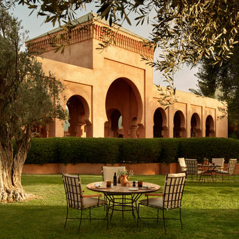 Olive grove at Amanjena with ochre pavilion, dining chairs and table on manicured lawn at sunset.