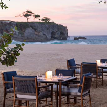 Beachclub dining tables set on sand at Amanera at sunset, with coastal cliffs beyond.
