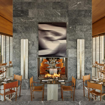 Amanzoe's main dining venue with wooden chairs, stone columns, and floor-to-ceiling windows overlooking the landscape.