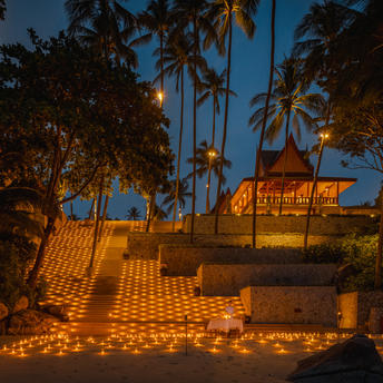 アマンプリのリゾートで、夕暮れ時に灯された火のランタンが地面を照らし、ヤシの木が暗い空に映える。