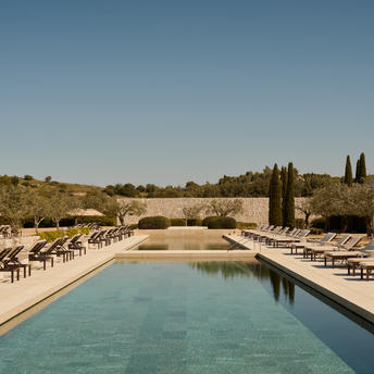 Rectangular pool with turquoise water reflecting clear sky, sun loungers lining the edges, cypress trees and stone architecture visible at Amanzoe resort.