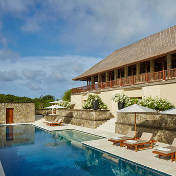 Infinity pool reflecting sky at Aman Villas at Nusa Dua, with thatched-roof villa and loungers beyond.