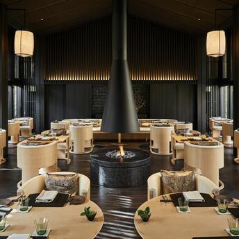Dining pavilion at Aman Kyoto with central fireplace and symmetrical table arrangements beneath wooden ceiling details.