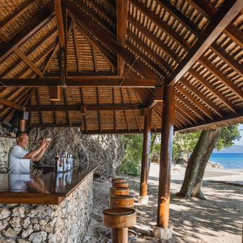 Beach bar at Amanwana with wooden pavilion, terracotta vessels, and views toward the shoreline.