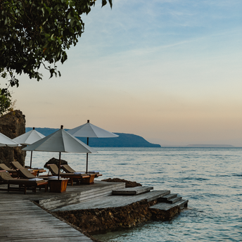 Wooden boardwalk at Amanwana with blue umbrellas and loungers overlooking calm waters at dusk.