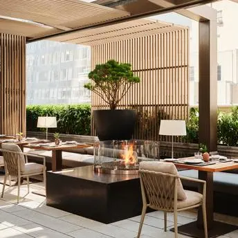 Terrasse de jardin avec salle à manger à l'Aman New York, hôtel avec tables en bois, plantes vertes et baies vitrées.