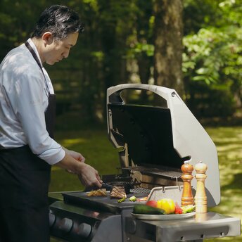 A staff member prepares food on a barbecue grill at Aman Kyoto, with fresh ingredients and greenery visible in the background.
