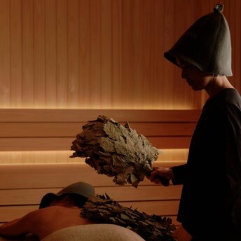 Person in traditional sauna attire at Aman Nai Lert Bangkok holding birch whisk over heated stones.