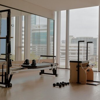 Bright gymnasium at Aman Nai Lert Bangkok with floor-to-ceiling windows overlooking the city skyline, exercise equipment and weights visible.