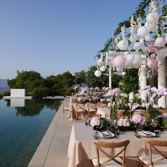 Outdoor dining terrace at Amanzoe overlooking calm waters, with flowering pergola and wooden chairs arranged for an event.