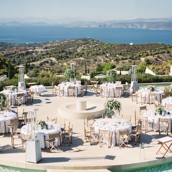 Seaside celebration venue at Amanzoe resort with dining tables and fire pit overlooking Greece's coastline.