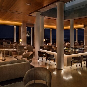 Amanzoe's dining and bar area illuminated at night, with contemporary seating, stone columns, and expansive windows overlooking the landscape.
