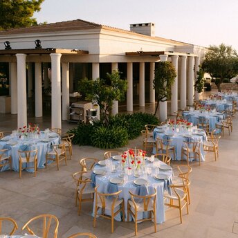 Wedding reception at Amanzoe resort with blue-dressed tables arranged on stone terrace beneath classical colonnade.