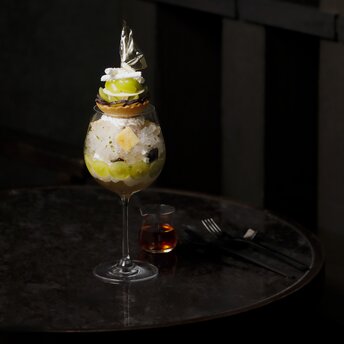 Shine Muscat cocktail at Aman Tokyo's lounge, served in a tall glass with garnish, beside a pair of sunglasses on a dark table.