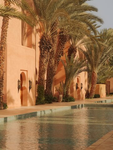 Amanjena's fountain courtyard with palm trees and terracotta walls reflected in still water.